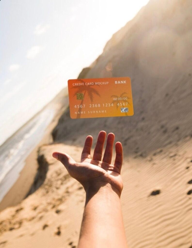 دست فردی در حال پرتاب کردن یک کارت اعتباری نارنجی در ساحل شنی زیر نور آفتاب